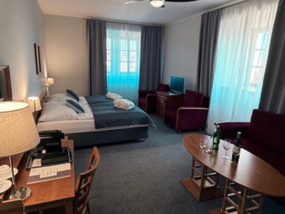 Hotel room with a double bed, blue bedding, two burgundy chairs, a desk, and a round table with wine on it