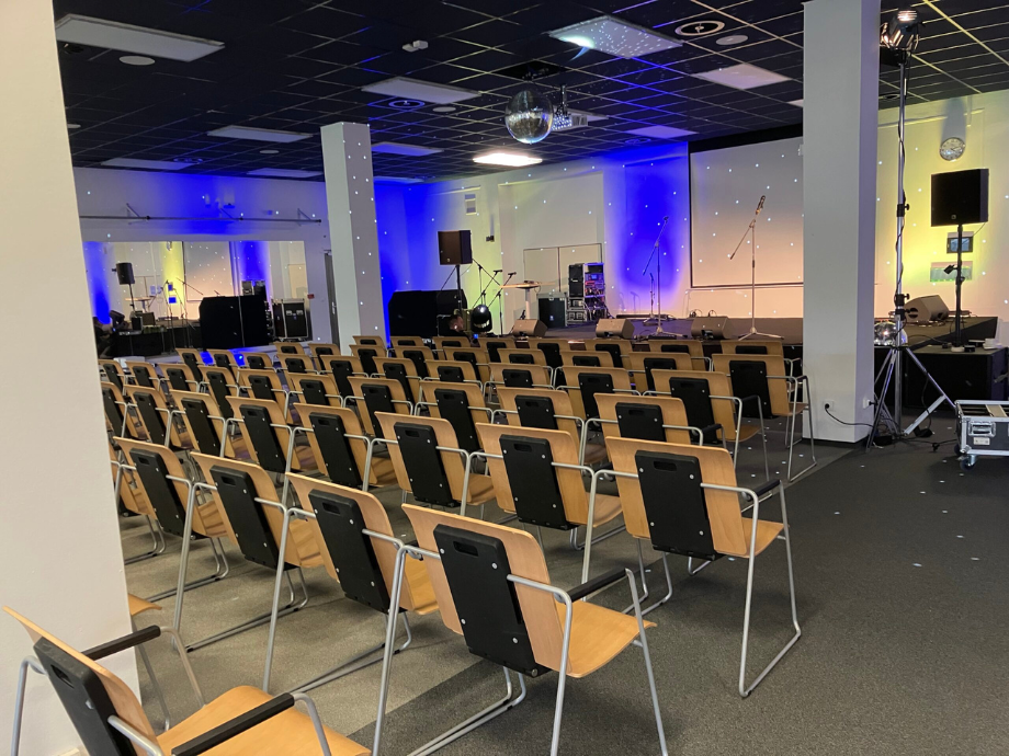Empty event hall with a small stage and microphones; rows of wooden chairs face the stage under blue wall lighting.