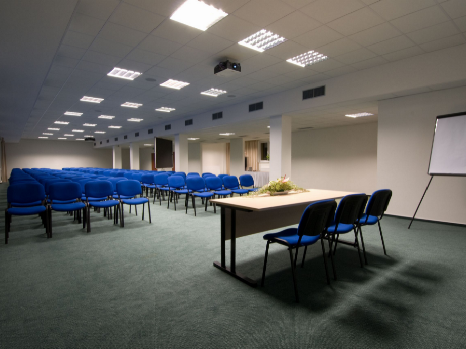 Conference room set up for a presentation: rows of blue chairs face a front table with flowers and presentation equipment (projector, whiteboard).