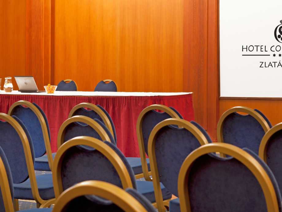 Empty hotel conference room with blue chairs facing a long table covered by a red skirt, laptop, pitcher, and fruit bowl against wood-paneled walls.