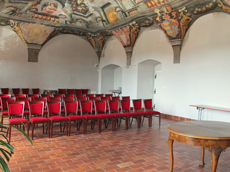 Rows of red-upholstered chairs set in a historic room with a painted vaulted ceiling and terracotta tile floor. A round wooden table sits to the right.