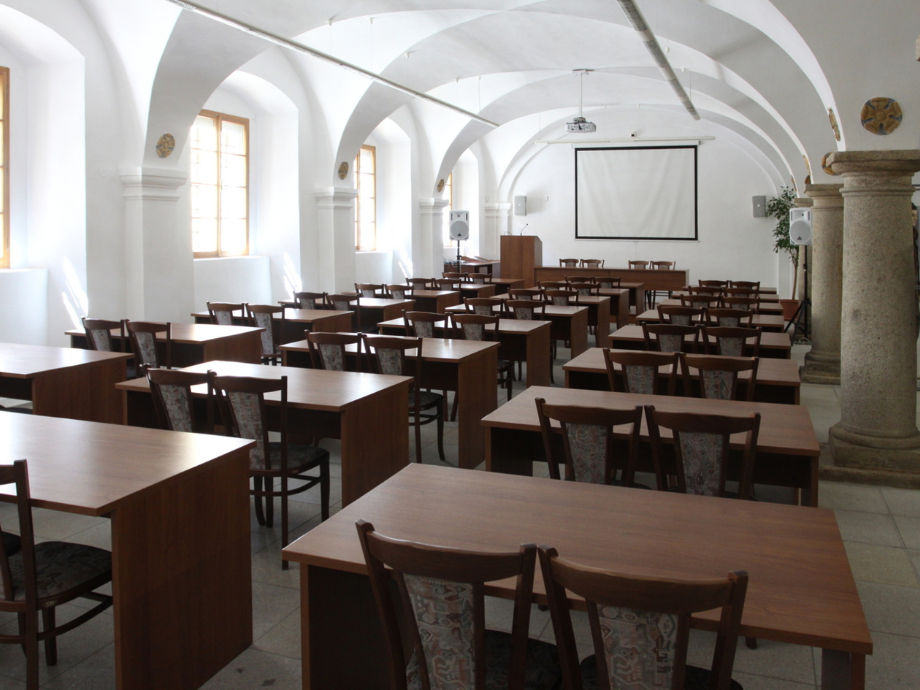 Lecture hall with rows of wooden desks and chairs facing a projector screen at the front.