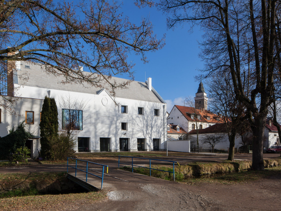 White two-story building with a gray roof, leafless trees, and a small blue railing bridge in the foreground under a blue sky