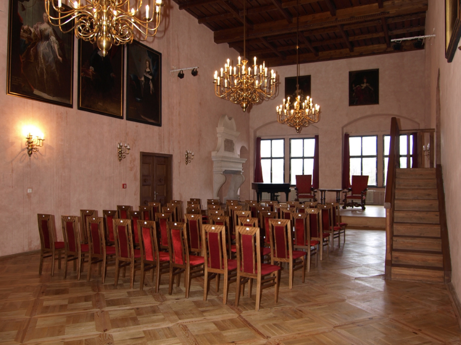 Elegant historic hall with red-upholstered chairs arranged in rows, chandeliers, and a stage with a piano at the far end.