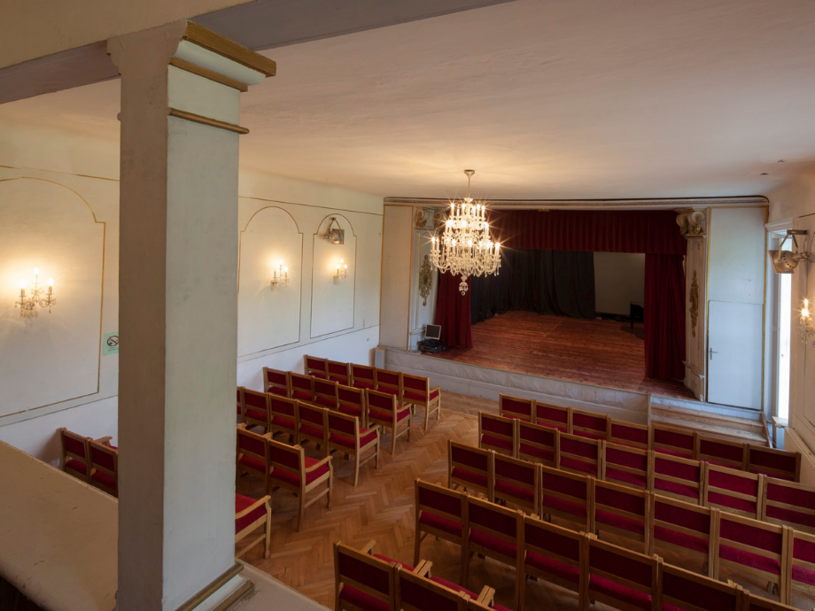 Empty historic theater with red upholstered seats facing a small stage, chandelier overhead, and a column in the foreground.