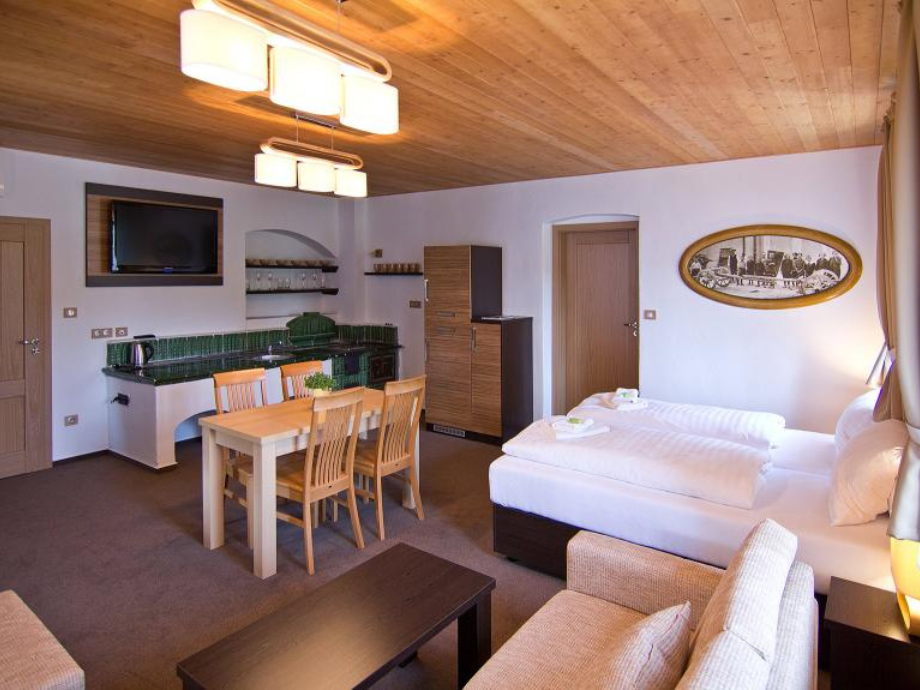 Hotel suite with two single beds, living area, and kitchenette under wood ceiling; TV on wall, dining table, and sofa arrangement visible