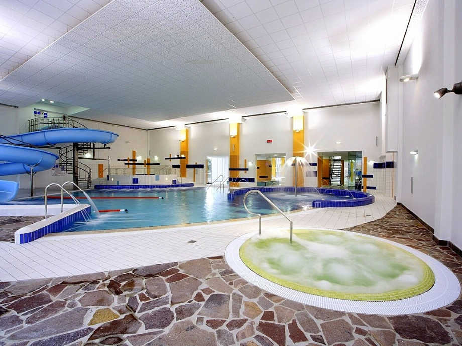 Indoor aquatic center: large pool with blue slides on the left, surrounded by tiled walls and bright lighting.