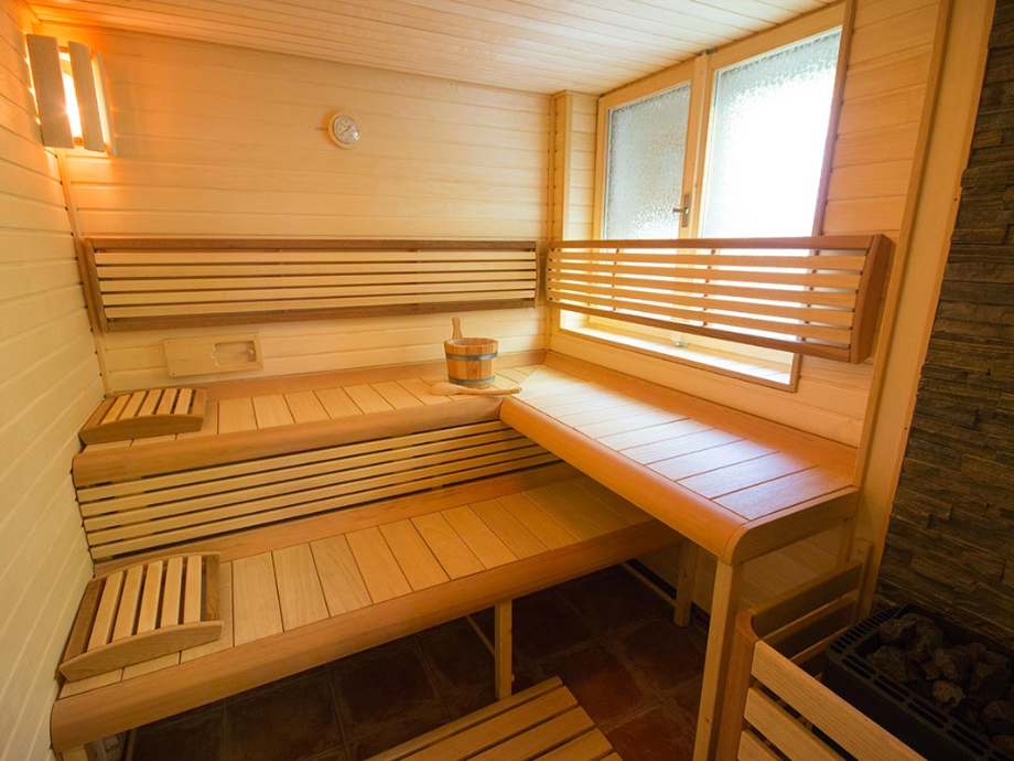 Wooden sauna interior with two-tier benches, a water bucket and ladle, and a stone heater in the corner.