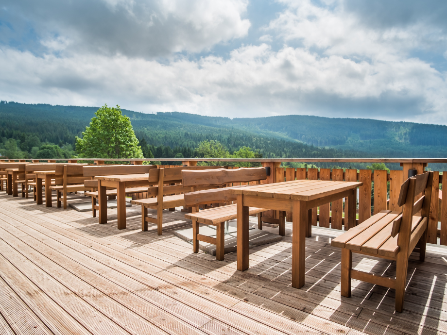 Wooden outdoor deck with tables and benches, overlooking a forested valley and distant mountains under a cloudy sky.