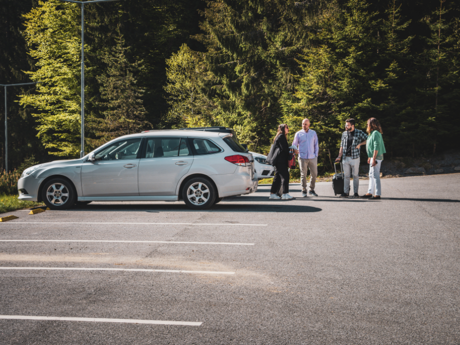 Five people with rolling suitcases stand beside a silver station wagon in a parking lot, with a forest backdrop.