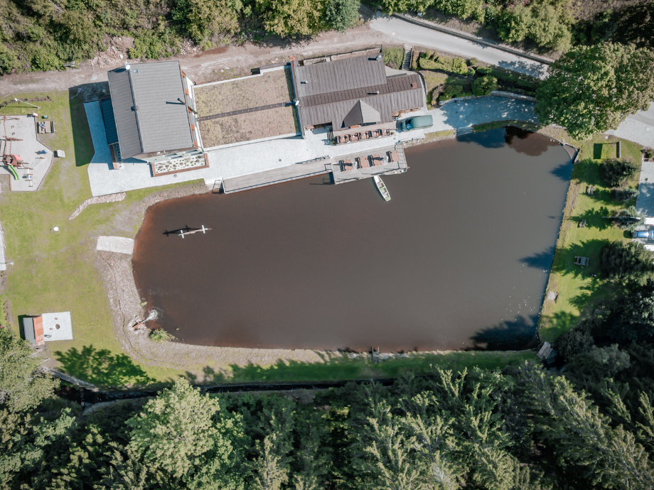 Aerial view of a rectangular pond with a wooden dock, two buildings on the shore, and surrounding trees and lawn.”