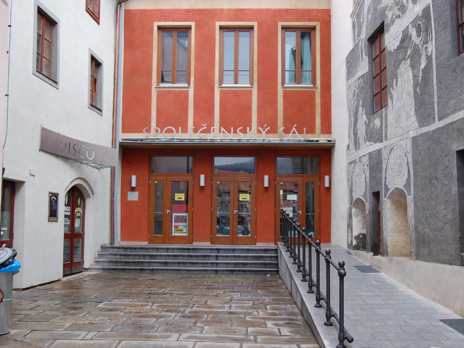 Facade of a cultural center with an orange facade, three tall windows, and 'SPOLEČENSKÝ SÁL' above glass doors; a ramp runs along the right and steps lead to the entrance.