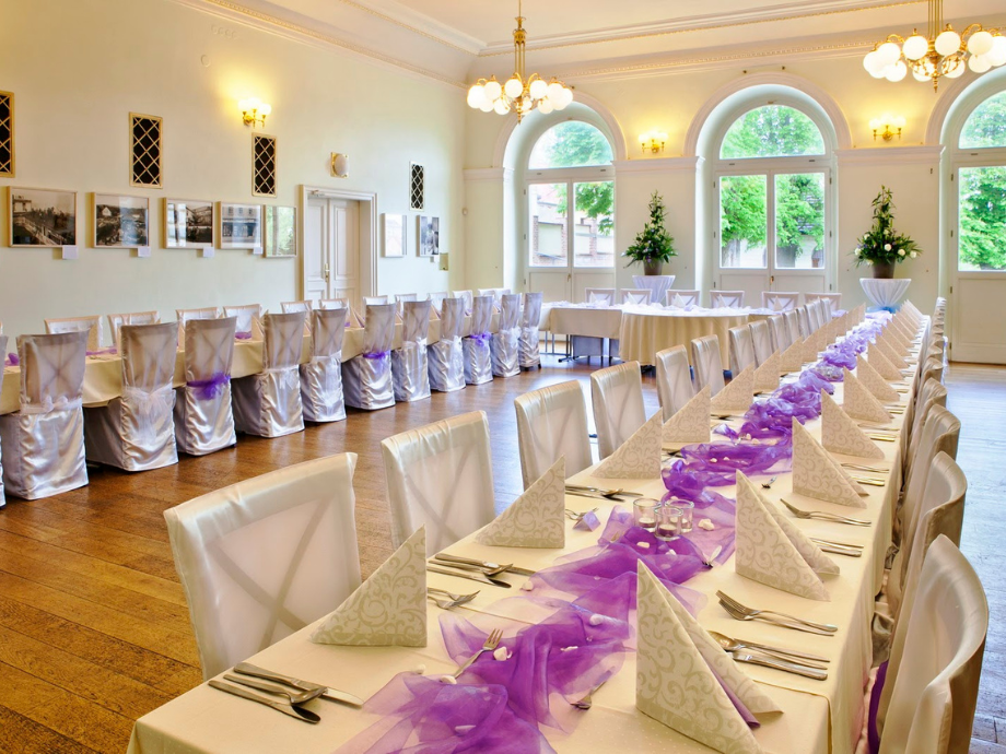 Elegant banquet hall arranged for a formal event with long tables, white chair covers, and purple fabric accents near large arched windows.