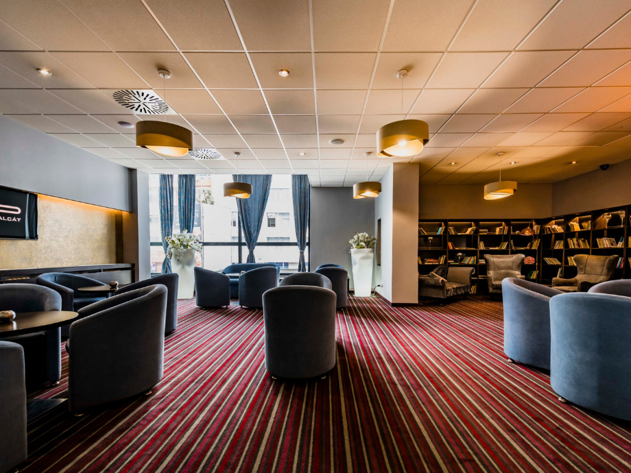 Spacious lounge with blue upholstered chairs, round tables, and a striped burgundy carpet; bookshelves line the right wall and large windows with blue curtains invite daylight.