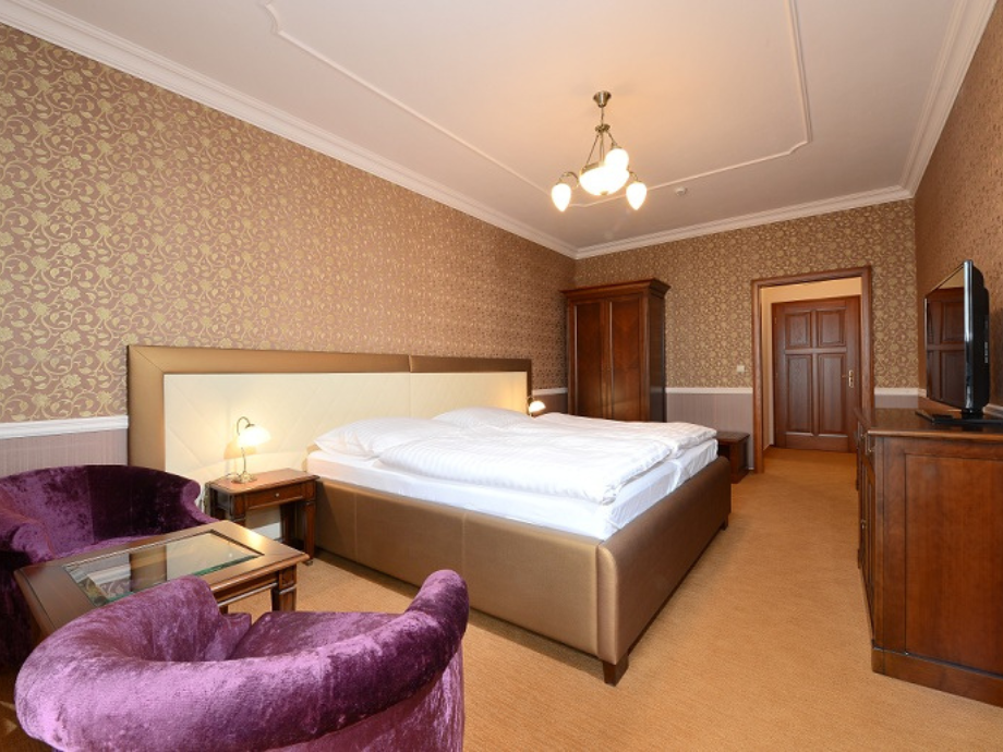 Hotel room with a king-size bed, two purple velvet chairs, and wooden wardrobe against ornate wallpaper.