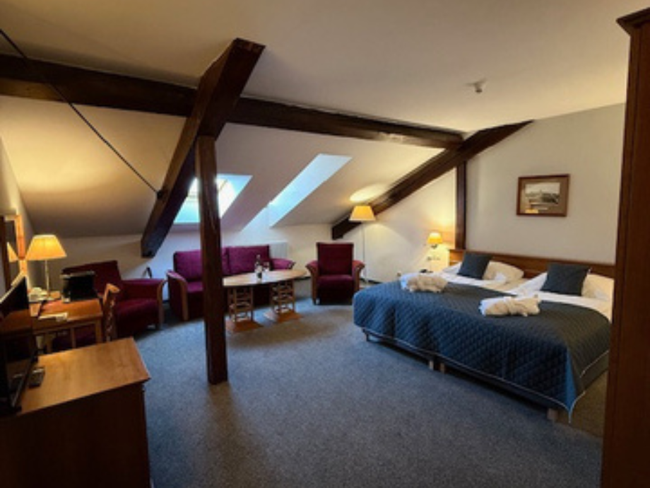 Loft-style hotel room with exposed dark wooden beams, a king-size bed with a blue quilt and folded towels, and a seating area with red chairs and a sofa.