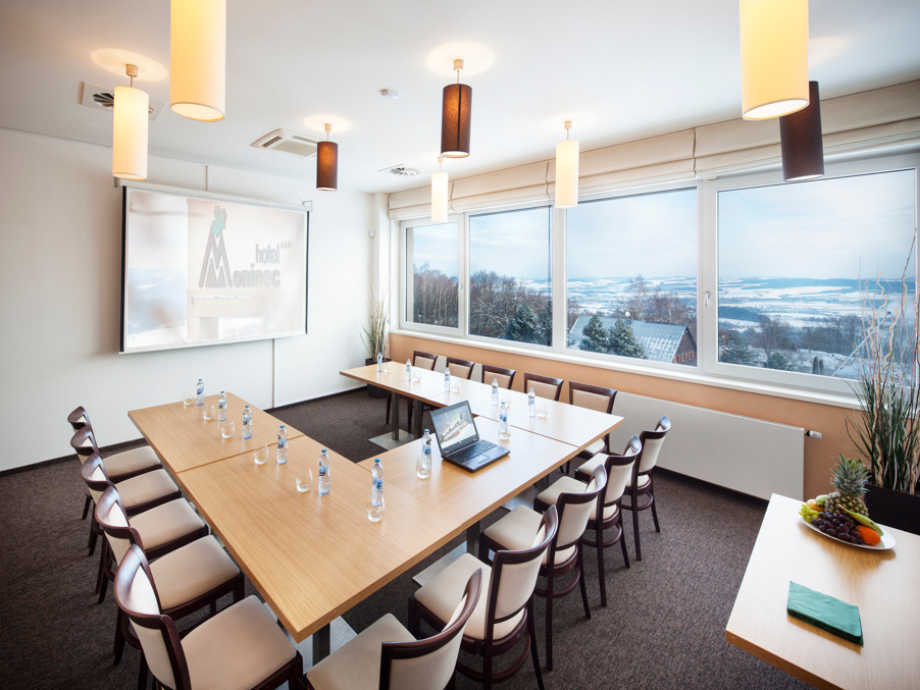 Conference room with a U-shaped wooden table, chairs, water bottles, a laptop, and a projector screen; large windows overlook a snowy landscape.