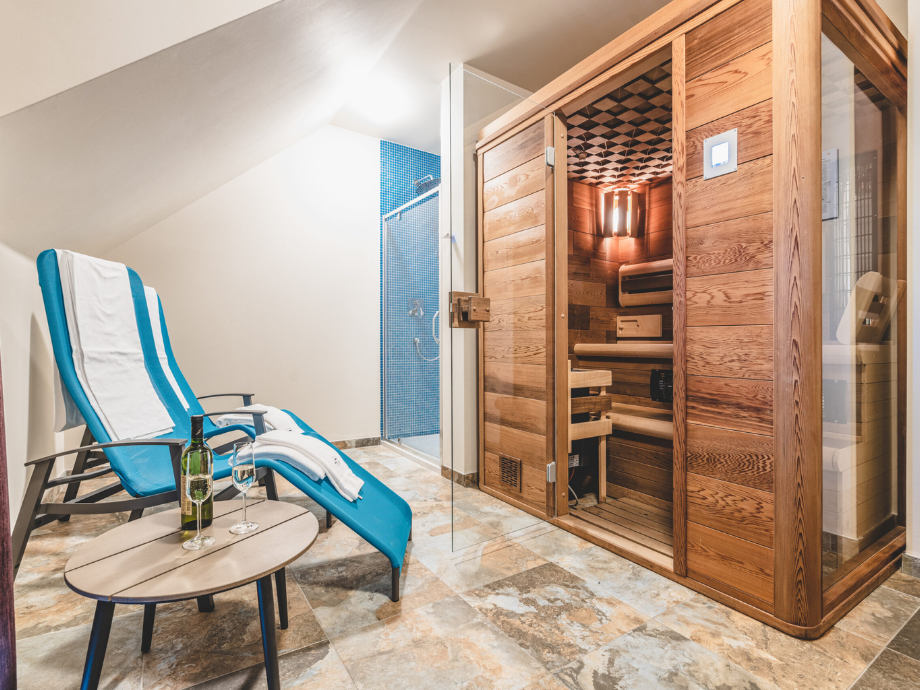 Spa-like room with a blue chaise lounge, small table with a wine bottle and two glasses, and a wooden sauna to the right; shower with blue tiles in back.