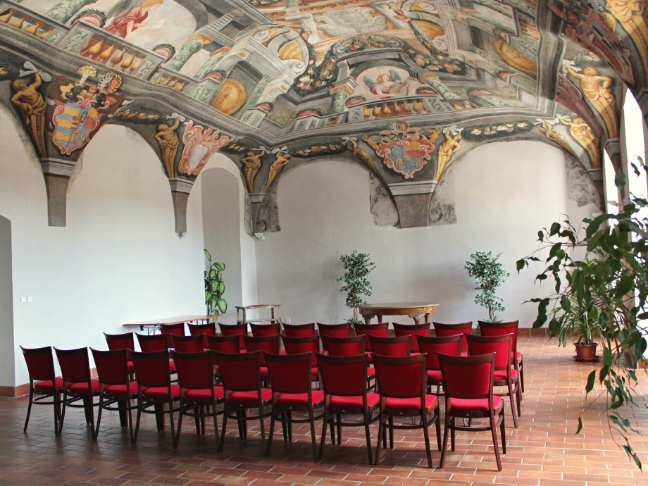 Rows of red chairs arranged in front of a frescoed, vaulted ceiling in a historic room with potted plants along the walls.