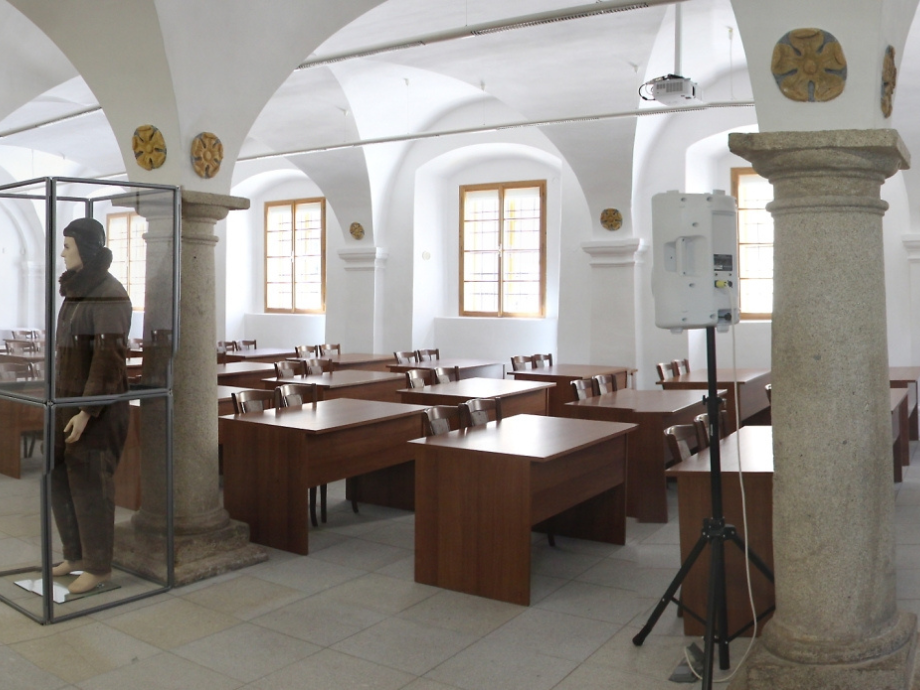A large classroom or lecture hall with wooden desks and stone columns, a glass display case containing a cloaked figure, and a projector on a stand.