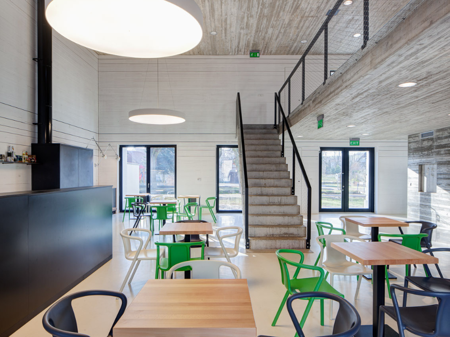 Modern cafe-like dining area with wooden tables, green and white chairs, a central concrete staircase, and large circular ceiling lights.