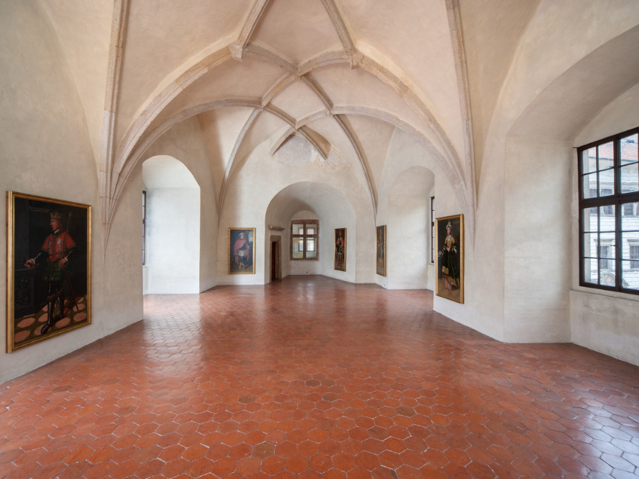 Interior of a historic gallery with arched vaulted ceiling, white walls, and paintings along the walls; red tiled floor.