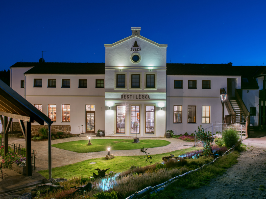 Nighttime exterior of a white distillery called Destilérka, warmly lit with a central entrance and manicured garden.