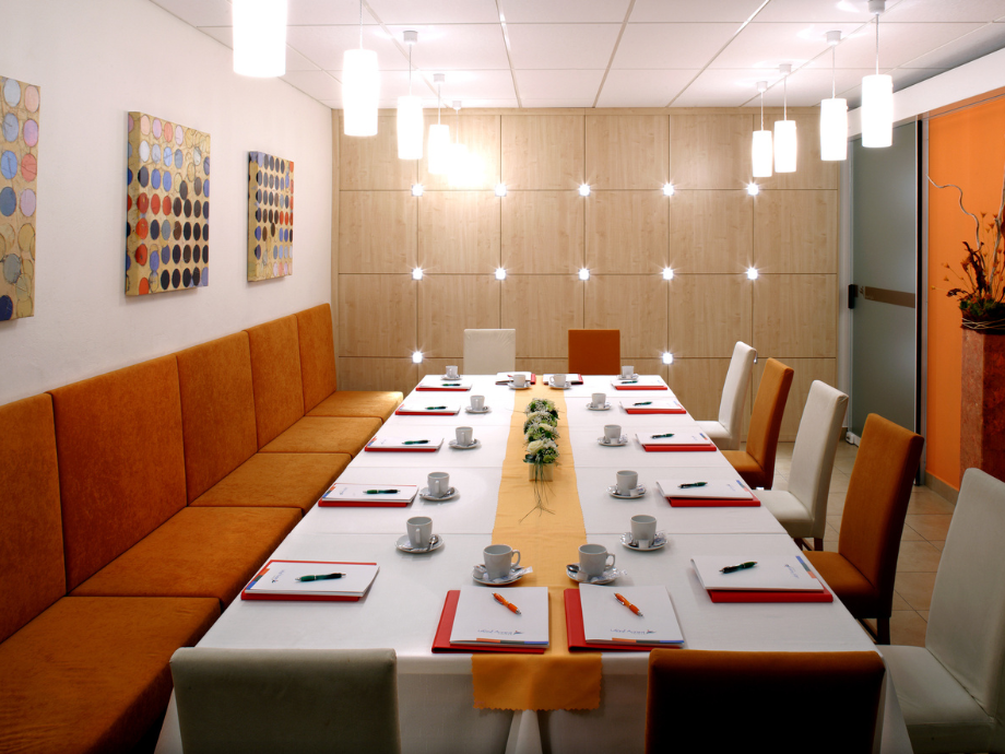 Long conference table set for a formal meal in a modern room, with orange upholstered benches, place settings, cups, and a yellow center runner.