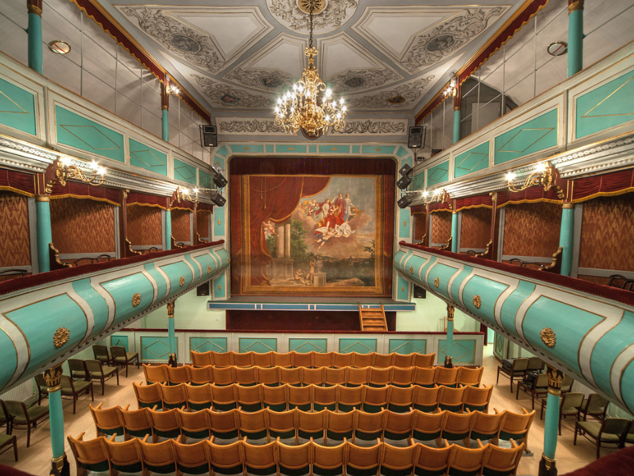 Grand, ornate theater interior with turquoise balconies, wooden seats facing a painted stage and chandelier above.