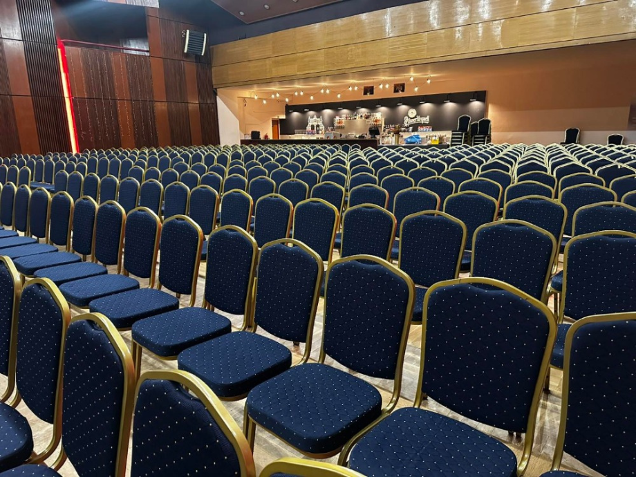 Large conference hall filled with rows of blue cushioned chairs with gold frames facing a stage/bar area in the distance.