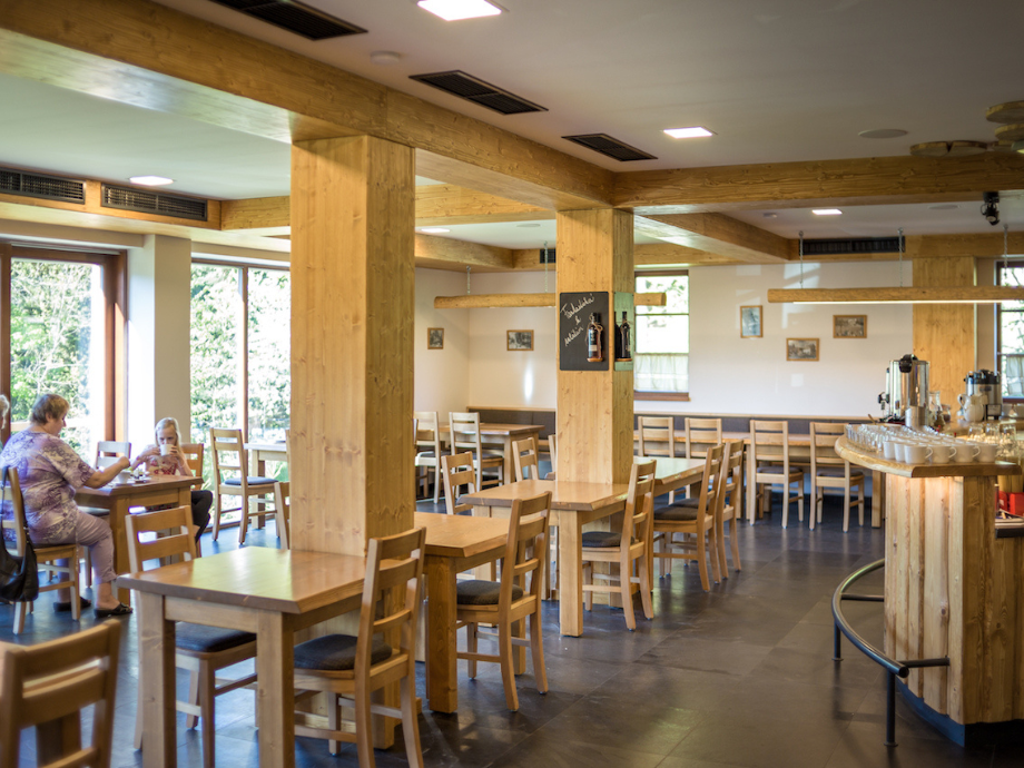 Cozy cafe interior with wooden beams, pillars, and rows of tables and chairs; a curved bar area on the right. Two customers sit by a window.