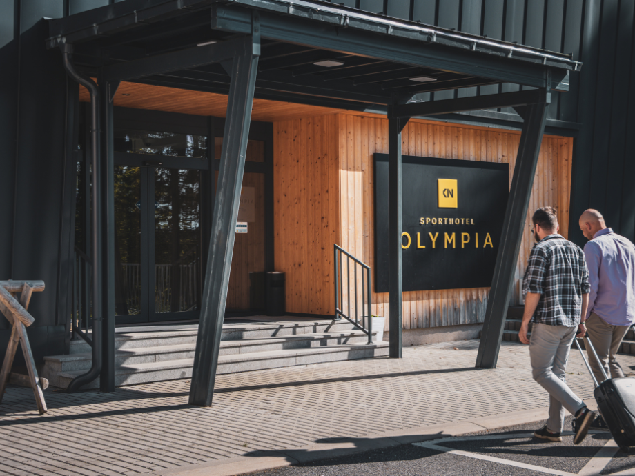 Hotel entrance with a wooden facade and a large black sign that reads 'Sporthotel Olympia'; two men walk toward the door with a rolling suitcase on a sunny day.