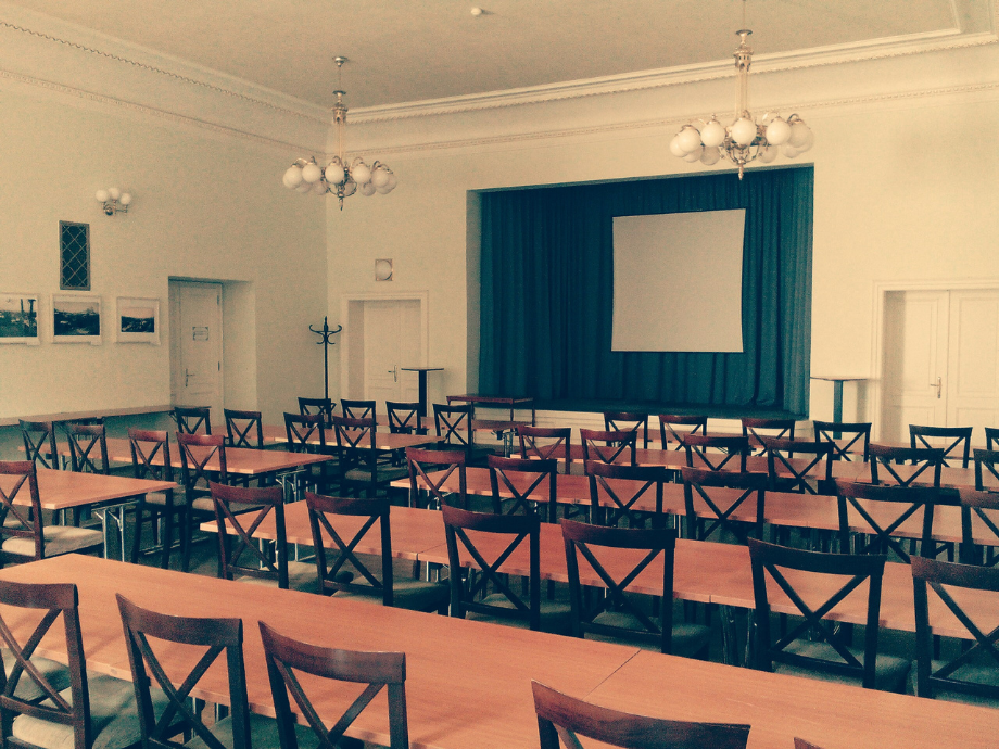 A classroom-style event hall with wooden chairs and tables facing a stage with blue curtains and a blank screen.