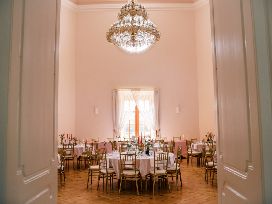 Elegant banquet hall with a crystal chandelier overhead, round tables with white linens and gold chairs set for a formal event.