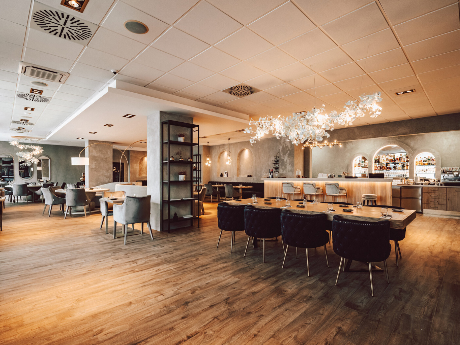 Open, upscale restaurant dining area with wooden floors, round and long tables, and a lit bar in the background in warm lighting.