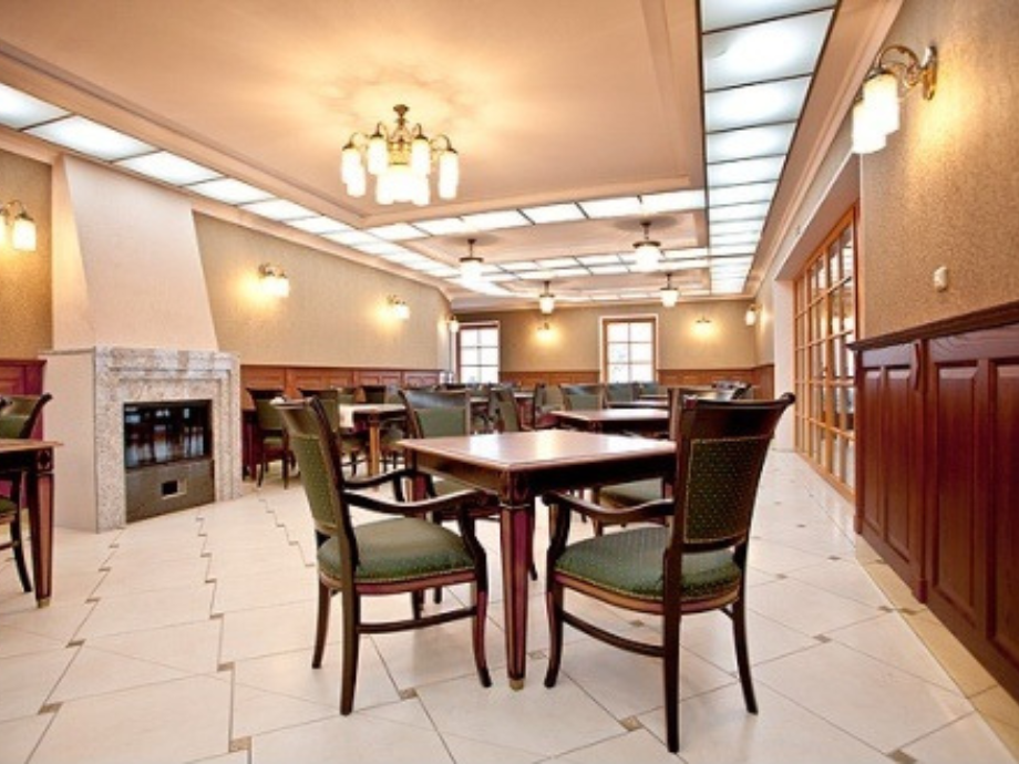 Interior of a restaurant dining area with square wooden tables, green upholstered chairs, and warm ceiling lights. A stone fireplace is visible on the left.