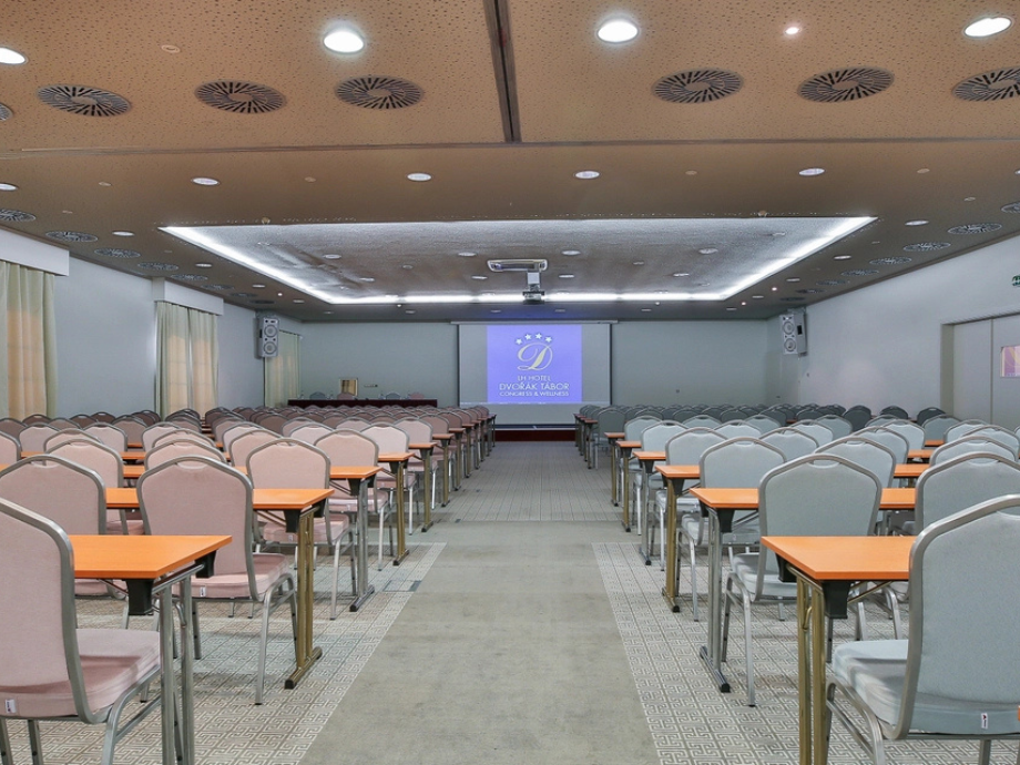 Large conference room set up for a presentation with rows of desks and cushioned chairs facing a projection screen at the front.