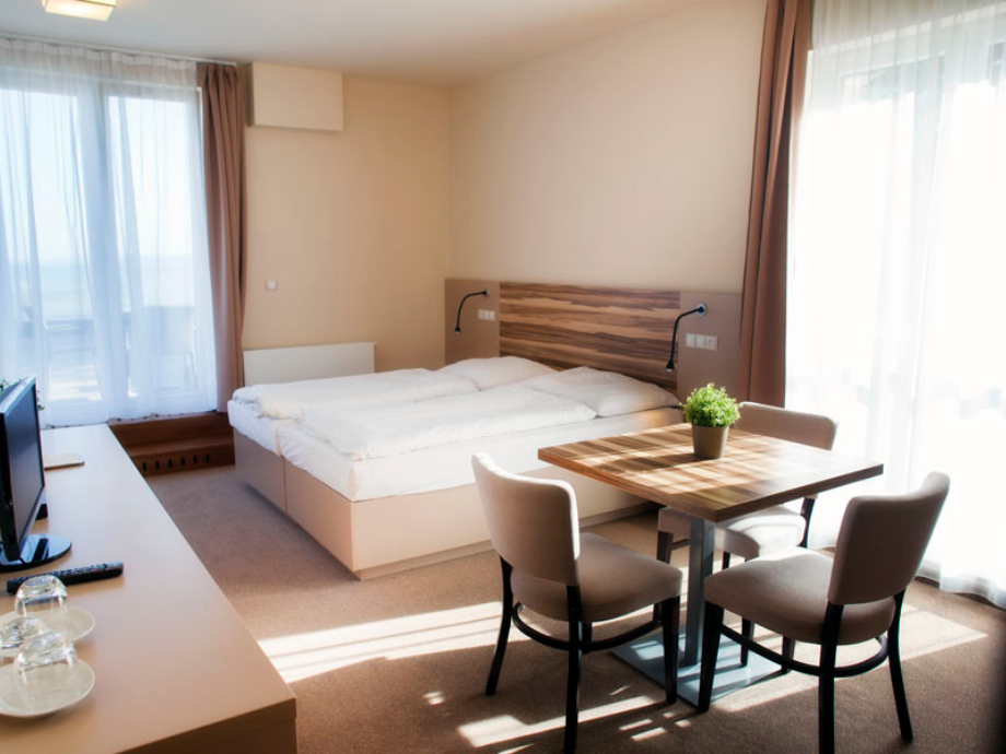 Bright hotel room with a double bed, wooden headboard, and a small dining table with a potted plant and three beige chairs by large windows with sheer curtains.