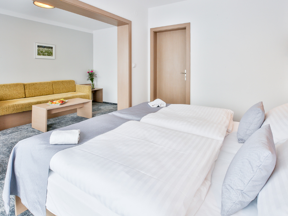 Two neatly made beds with white linens and gray throws overlook a living area with a mustard sofa, coffee table, and a small plant in a bright hotel room.