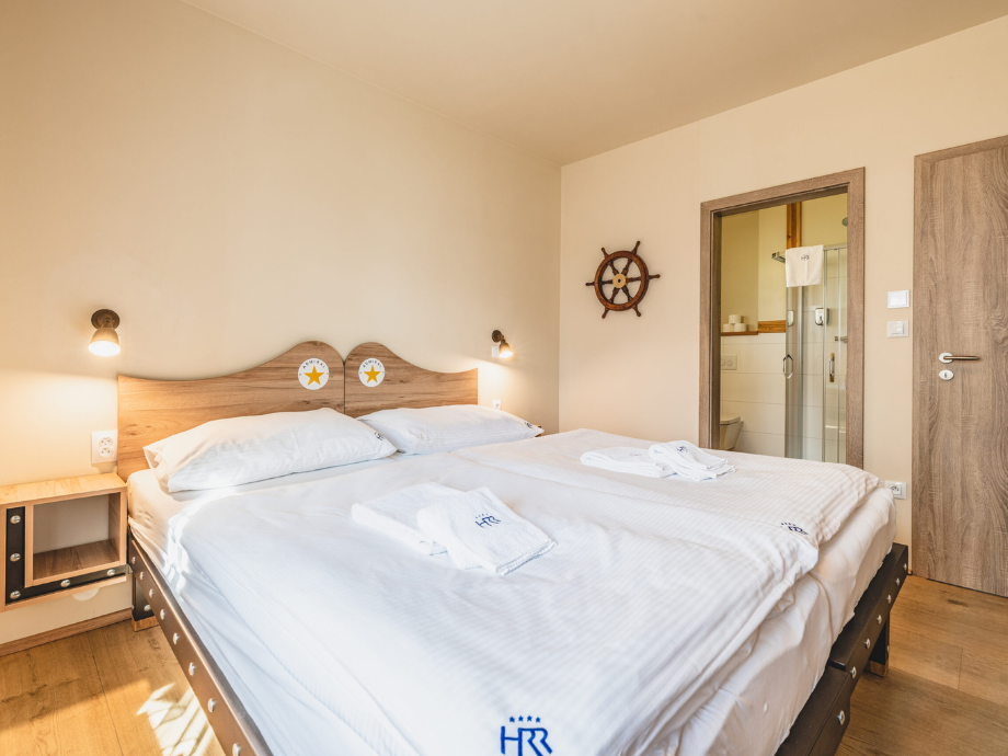 Hotel room with a large double bed, white linens and folded towels, wooden headboard, and bedside lamps; a ship wheel decor and a doorway to a glass-walled bathroom visible.