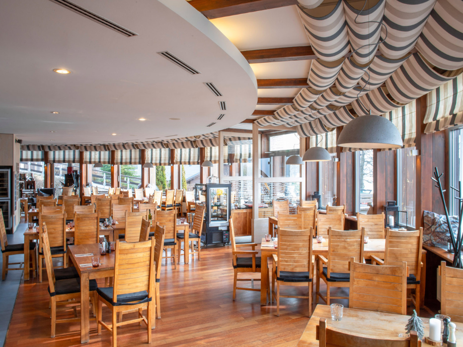 Wooden dining tables and chairs fill a bright restaurant with striped awning-style ceiling and large windows, creating a warm, open space.
