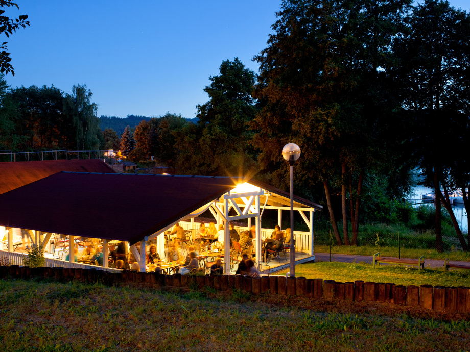 Outdoor dining pavilion with a dark red roof, warm lights, and groups of people eating at tables by the lake at dusk.