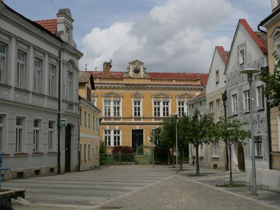 Historic town square with a central yellow-orange facade and decorative clock, bordered by white and pastel houses and a cobblestone street.