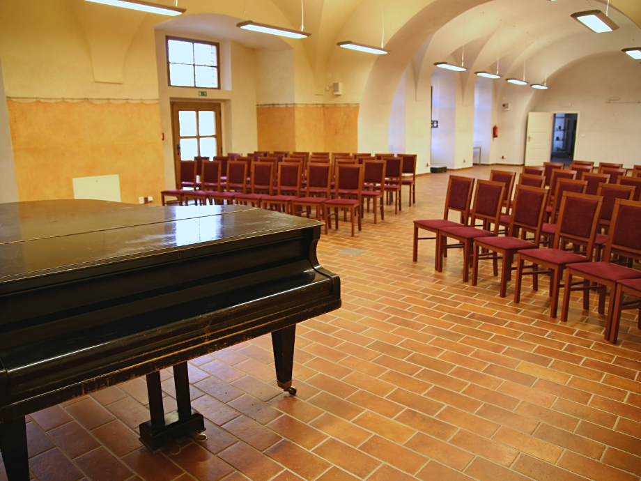 Black grand piano in a brick-floored recital hall with rows of red chairs and arched ceiling lighting in the background, empty.