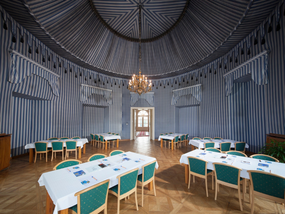 Banquet hall with blue-and-white striped walls, a crystal chandelier, and arranged tables covered in white cloths with green chairs.
