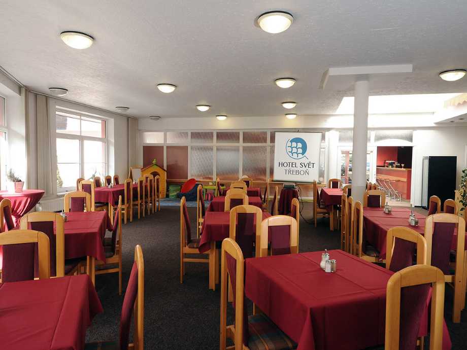 Hotel dining hall with red-cloth tables and wooden chairs, windows on left, and a sign reading 'Hotel Svět Třeboň' in the background.