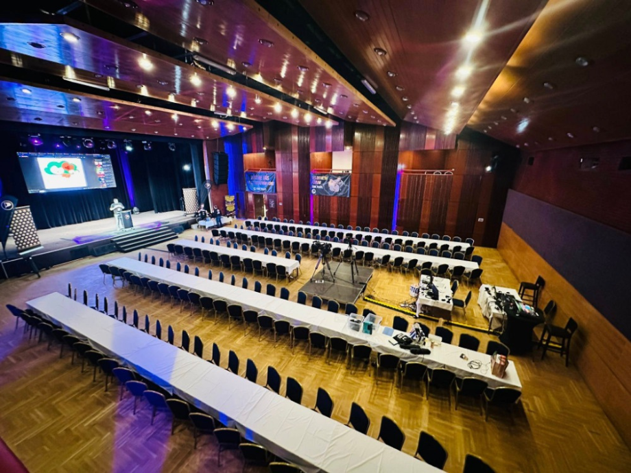Conference hall prepared for a presentation: long white-clothed tables with chairs facing a stage and large screen on the left.