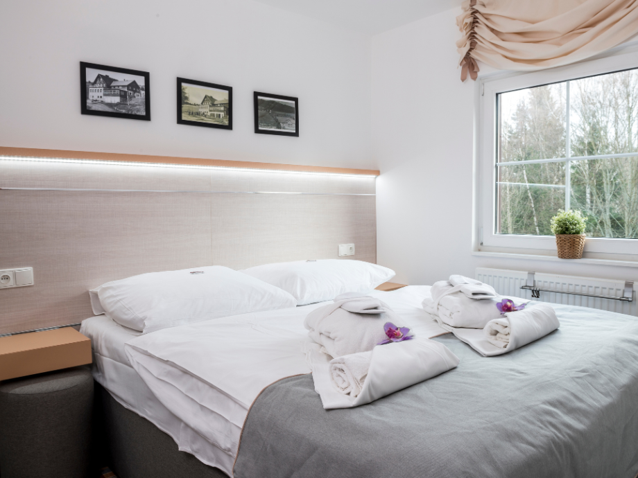 Bright hotel room with a neatly made bed, two pillows, and folded towels topped with purple orchids on a gray bedspread.