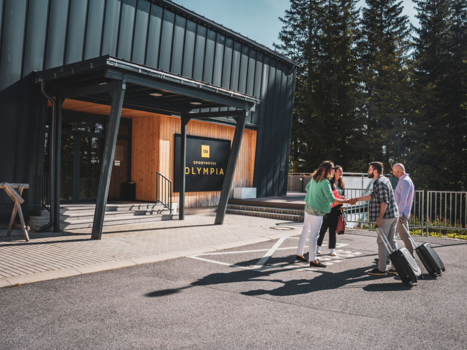 Group of four adults with rolling suitcases greeting each other outside a modern hotel entrance labeled Olympia Sporthotel Olympia Office or lobby area.