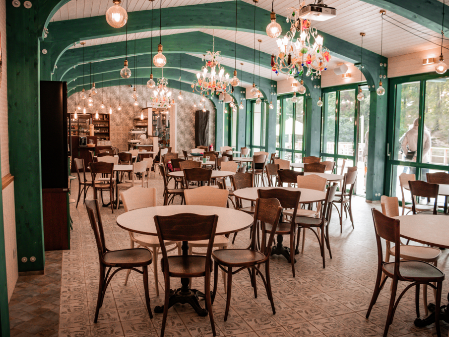 Bright cafe interior with round tables and wooden chairs, green arched beams, and decorative chandeliers.